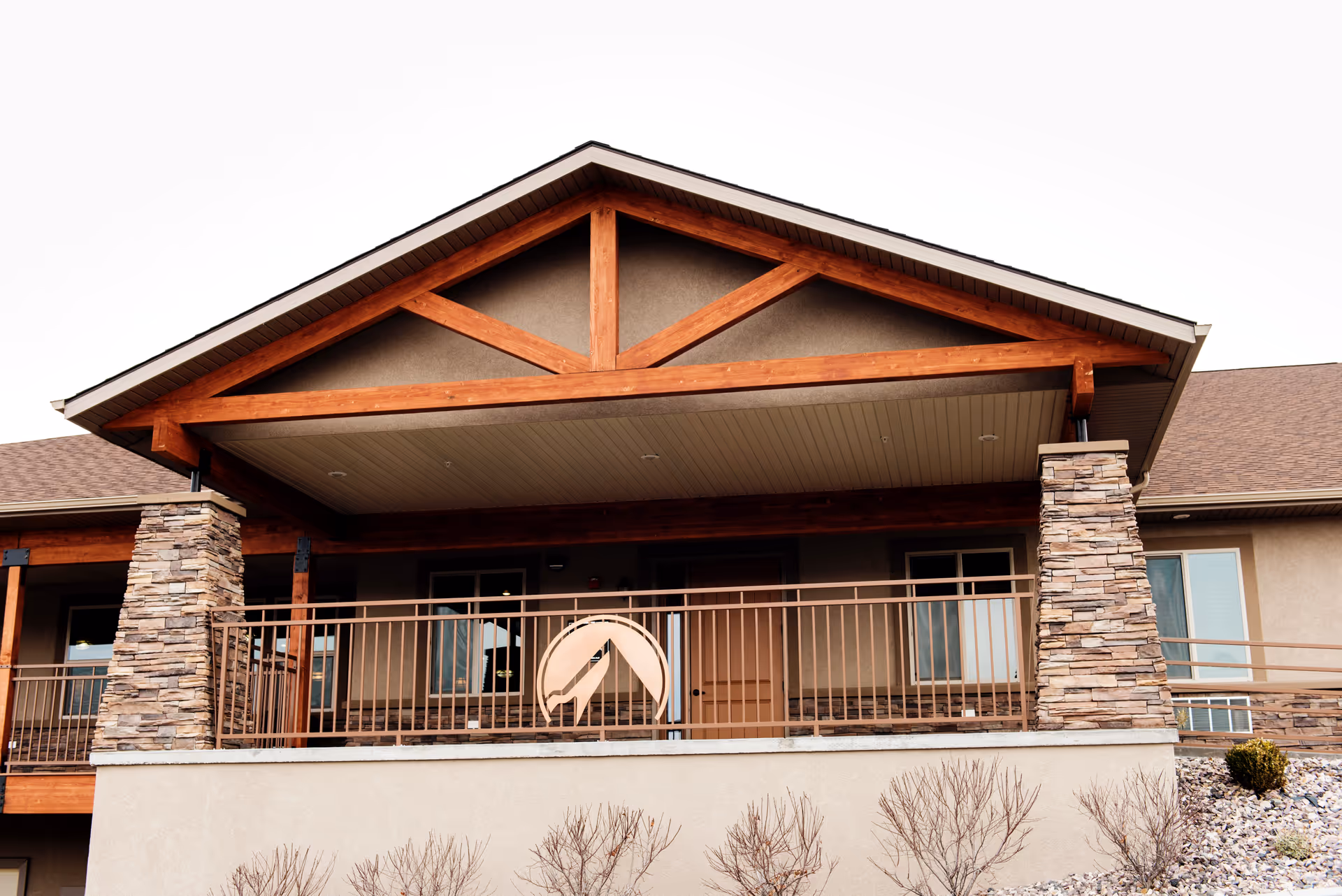 Front exterior view of a building with a covered porch supported by stone pillars and wooden beams. The porch has a metal railing with a circular emblem featuring a mountain design. The building has beige walls and a brown shingled roof.