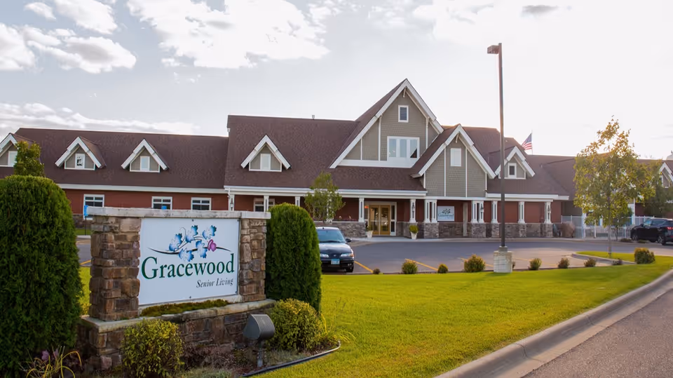 Exterior view of a senior living facility building with a large sign in front that reads 'Gracewood Senior Living'. The building has a pitched roof with multiple dormer windows, a stone facade at the base, and a parking lot with a few cars. The lawn is well-maintained with bushes and trees, and the sky is partly cloudy.