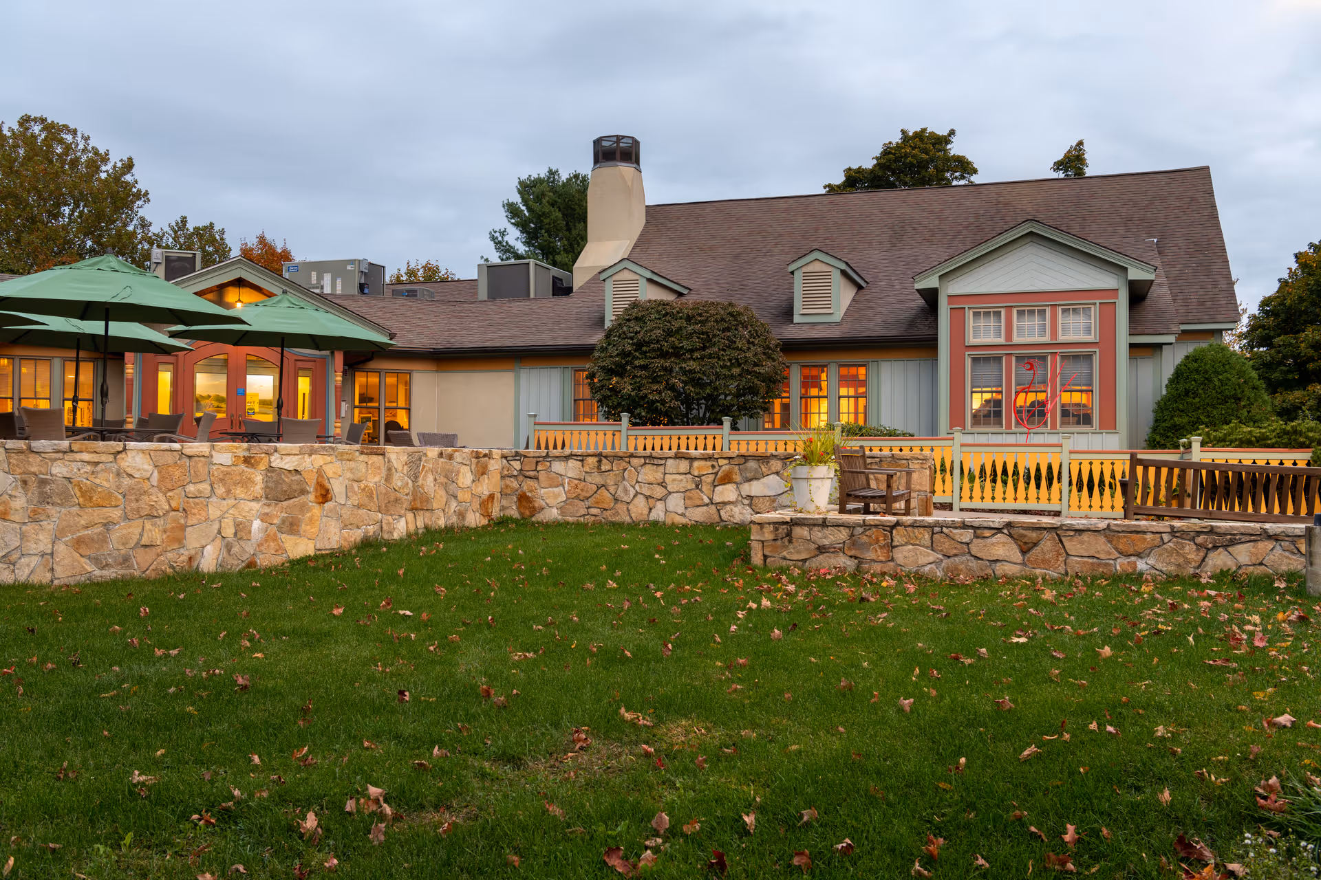 Exterior front view of a single-story senior living building with a stone wall, grassy lawn, patio umbrellas, and lit windows at dusk.