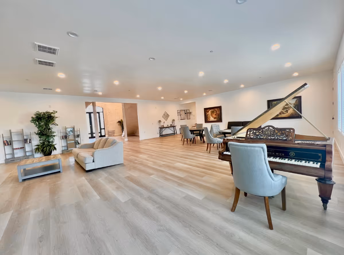 A spacious, well-lit common area in a senior living facility featuring a grand piano with a chair, several dining tables with chairs, a beige couch, a coffee table, bookshelves, and decorative plants. The room has light wood flooring, white walls, and recessed ceiling lights.