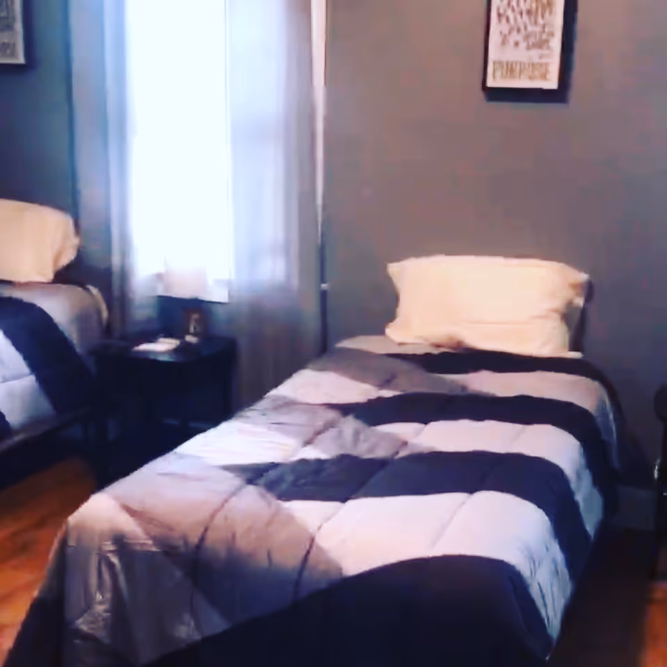 A bedroom with two single beds, each with a pillow and a striped comforter. There is a window with sheer curtains between the beds, and a small nightstand with a lamp and some items on it. The walls are painted gray and have framed artwork hanging.