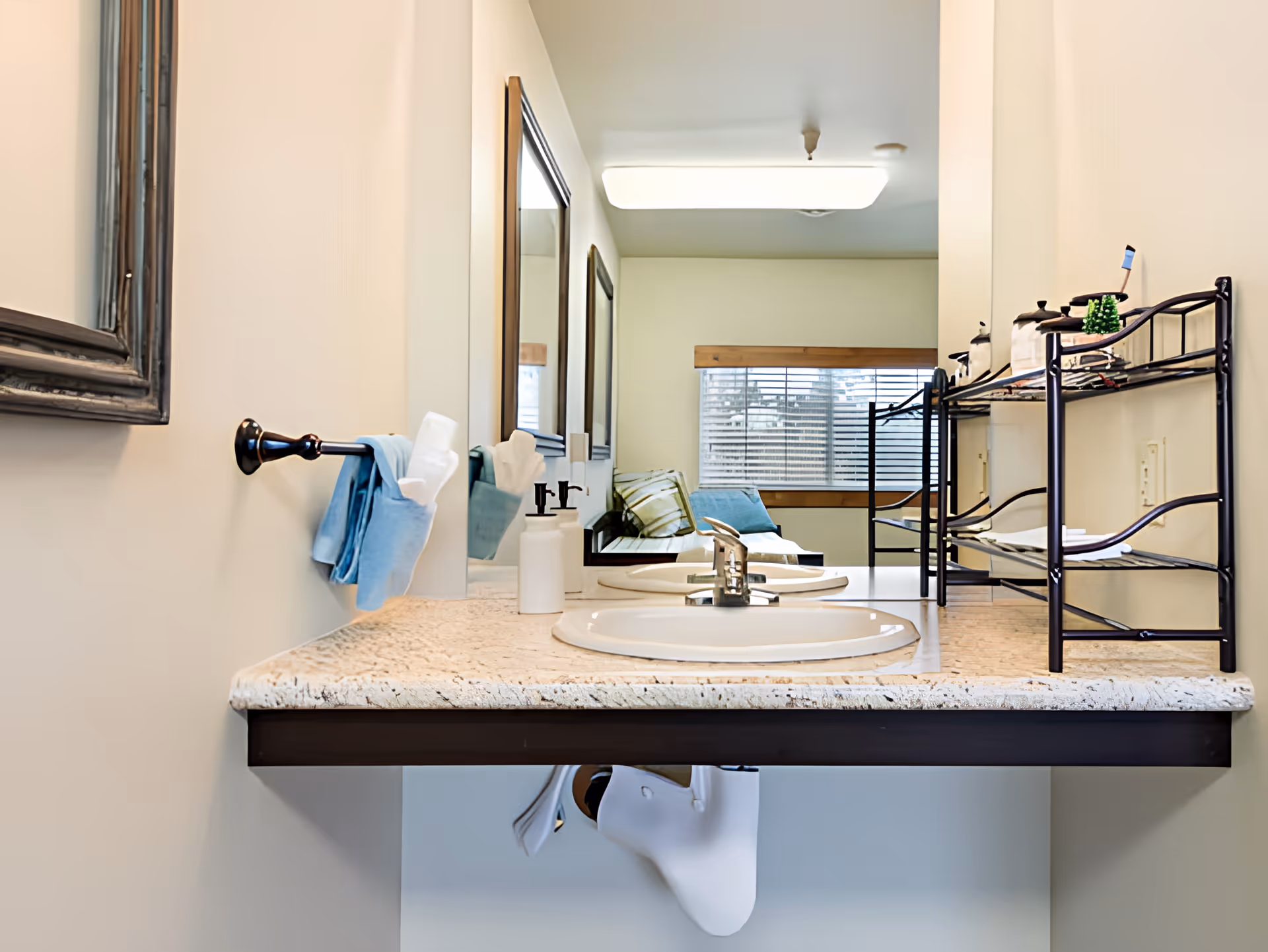 A bathroom vanity area with a single sink, a granite countertop, a mounted mirror, and a towel rack holding a blue towel and tissue. In the background, there is a window with blinds and a seating area with cushions visible through the reflection in the mirror.