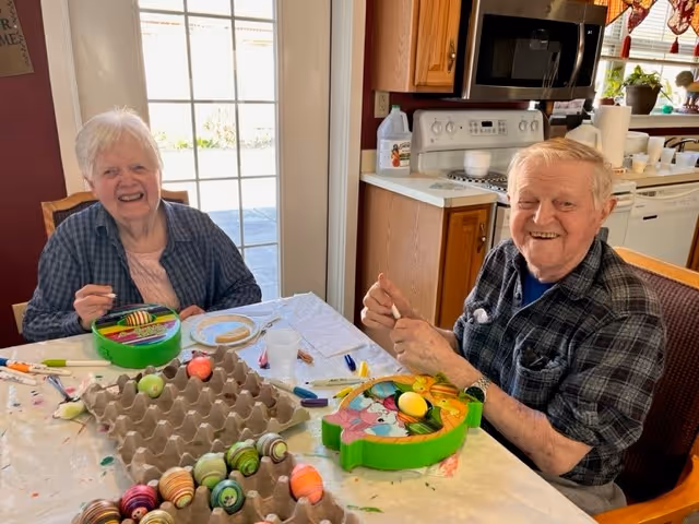 Two elderly people sitting at a table in a kitchen, smiling and painting colorful Easter eggs. The table is covered with a white cloth and has egg cartons and paint supplies scattered on it. The kitchen has wooden cabinets, a microwave, and a stove in the background.