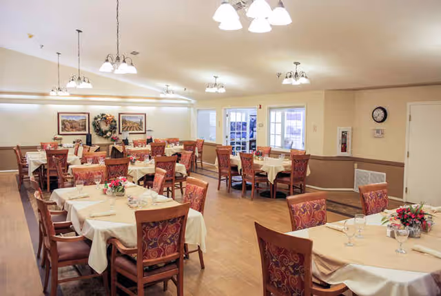 A dining room with multiple tables covered in beige tablecloths, each set with glasses, plates, and floral centerpieces. The room has wooden chairs with patterned upholstery, light-colored walls, framed artwork, and several ceiling light fixtures. There are windows and a door visible in the background.