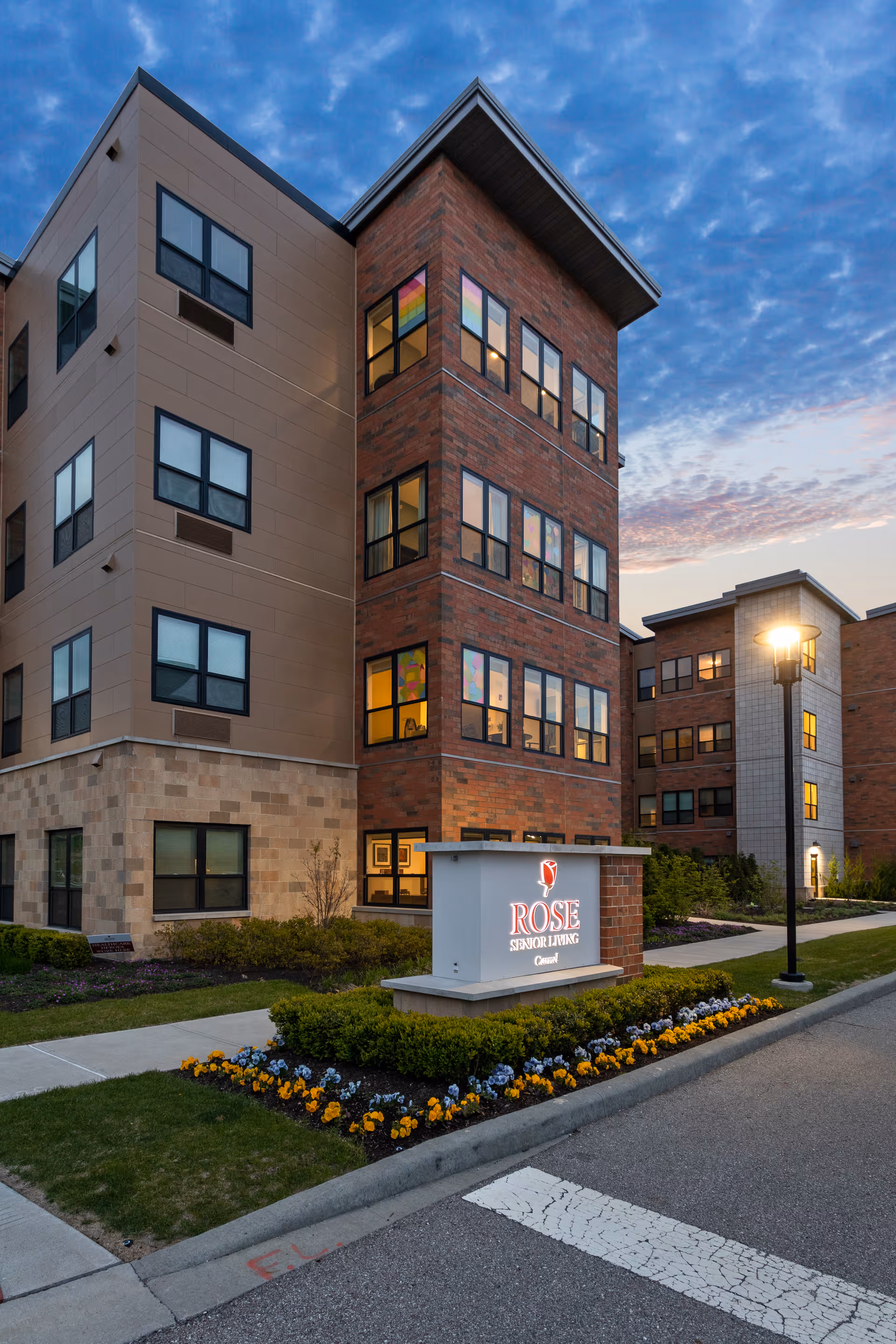 Exterior front view of a multi-story brick and stone senior living building at dusk with a lit 'ROSE Senior Living' sign and landscaped flower beds.