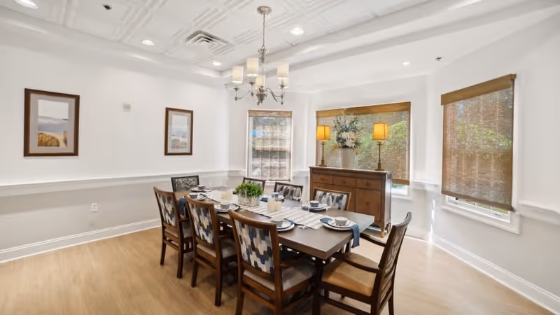 A bright dining room with a rectangular wooden table set for eight people. The chairs have patterned upholstery. There are three windows with woven shades, a wooden sideboard with two lamps and a floral arrangement, and two framed pictures on the wall. A chandelier hangs above the table.