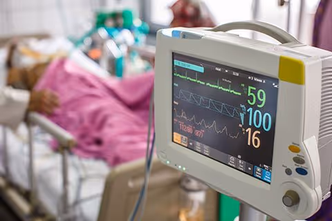 A medical vital signs monitor showing heart rate and oxygen levels in the foreground with a blurred patient lying in a bed under a pink blanket in the background.