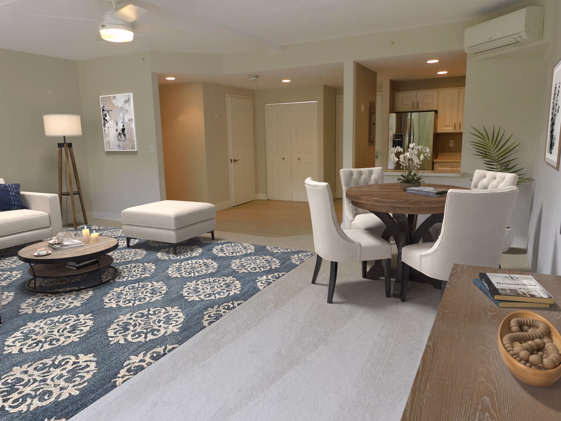 A bright and spacious living and dining area in a senior living facility. The living room features a beige sofa, a matching ottoman, a round wooden coffee table on a blue patterned rug, and a tall floor lamp. The dining area has a round wooden table surrounded by four upholstered chairs. The kitchen is visible in the background with white cabinets and stainless steel appliances. The walls are painted light beige, and there are decorative plants and framed artwork on the walls.