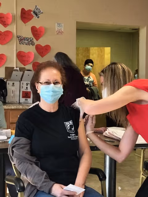 A woman wearing a face mask and glasses is seated and receiving a vaccination shot in her upper arm from a healthcare worker in a red shirt. The setting appears to be a healthcare or rehabilitation center with other people in the background also wearing masks. Red heart-shaped notes with messages are displayed on the wall behind them.