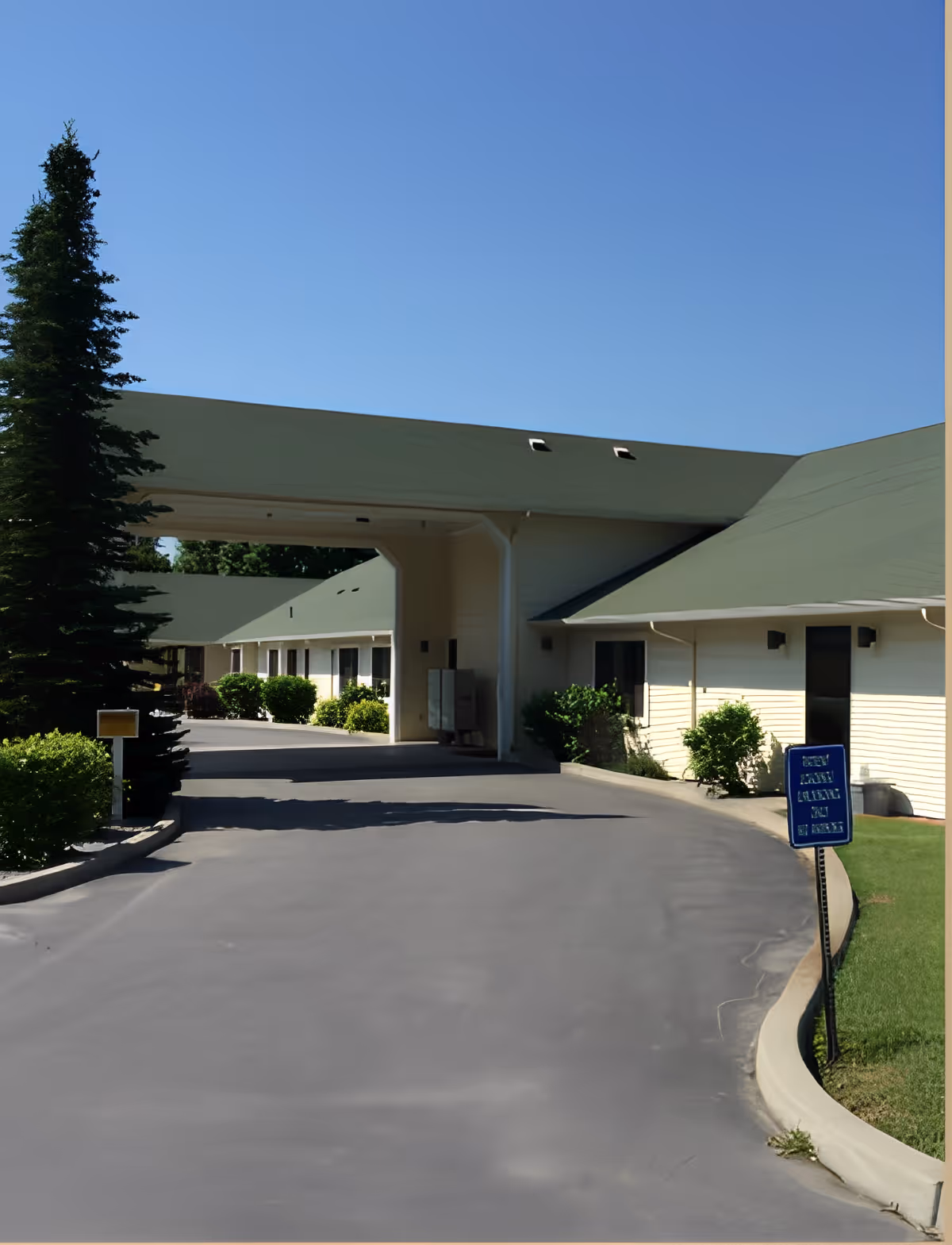 Exterior view of Buena Vista Healthcare facility showing a driveway leading under a covered entrance with green roofs and beige walls, surrounded by bushes and a tall tree under a clear blue sky.