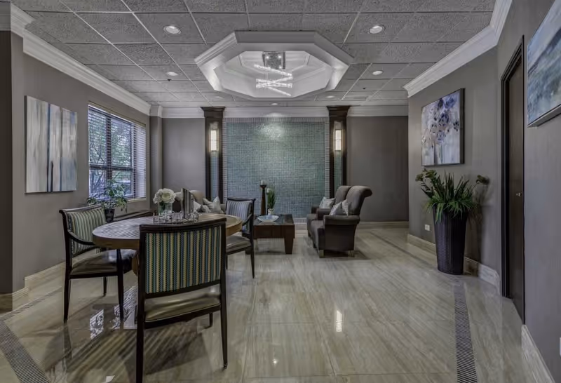 A modern and elegant common area with a round wooden table surrounded by four chairs with striped upholstery. There are two armchairs and a coffee table in the background against a wall with a decorative tiled panel and two vertical lights. The room has a tiled floor, gray walls, two paintings, a large window with blinds, and a ceiling light fixture with a geometric design.