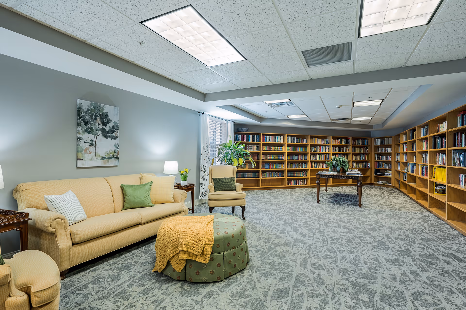A spacious and well-lit library room with beige and green upholstered seating including a sofa, armchair, and ottoman with a yellow throw blanket. The room features large wooden bookshelves filled with books along two walls, a wooden table with a plant on it, carpeted floor with a patterned design, and soft lighting from ceiling fixtures and table lamps. A painting of a tree hangs on the light gray wall above the sofa.