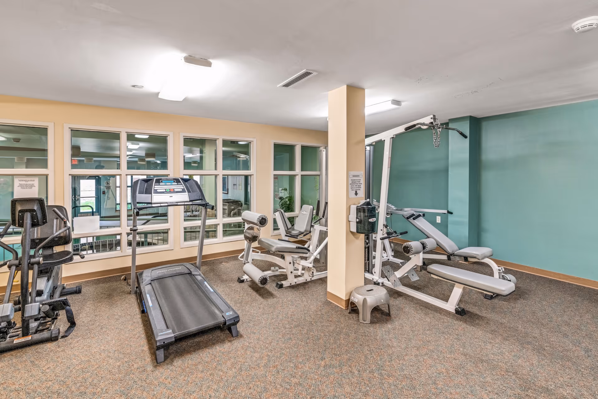 Small fitness room with a treadmill, elliptical, and weight machines under fluorescent lighting.