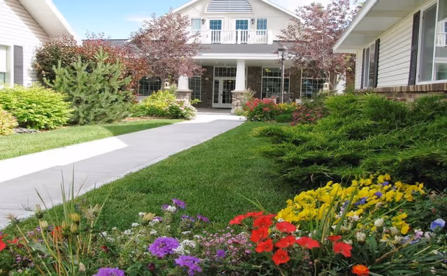 A well-maintained garden path leading to the entrance of a building with white siding and a balcony. The garden features colorful flowers and green shrubs on both sides of the concrete walkway under a clear blue sky.