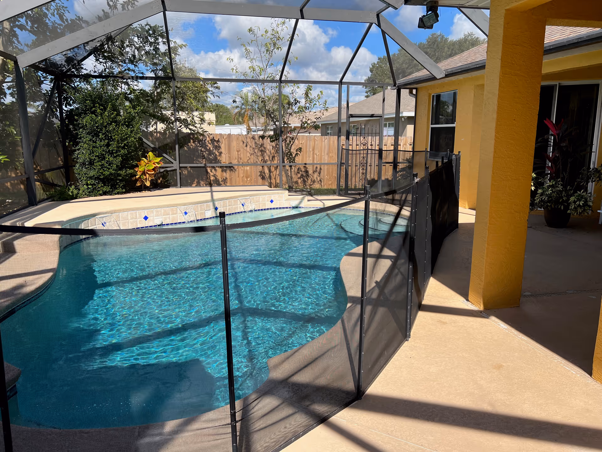 A screened-in outdoor swimming pool area with a safety fence around the pool. The pool is surrounded by a concrete patio and there are plants and trees visible outside the screened enclosure. The building adjacent to the pool is painted yellow with windows and a covered patio area.