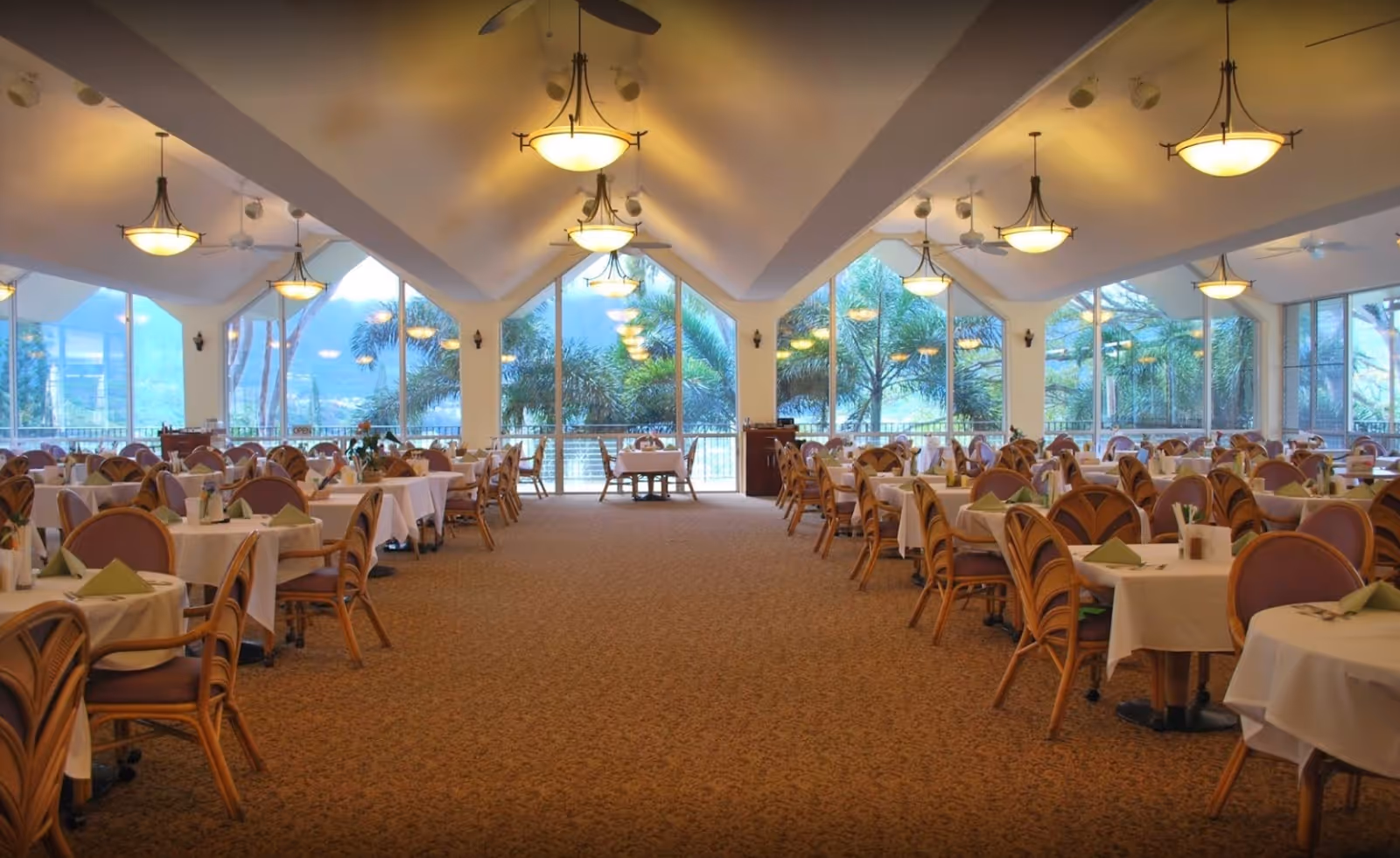 Spacious dining room with multiple tables covered in white tablecloths and set with green napkins and utensils. The room features large floor-to-ceiling windows with views of palm trees and mountains outside. The ceiling has multiple hanging light fixtures and ceiling fans, and the carpet is patterned in warm tones.