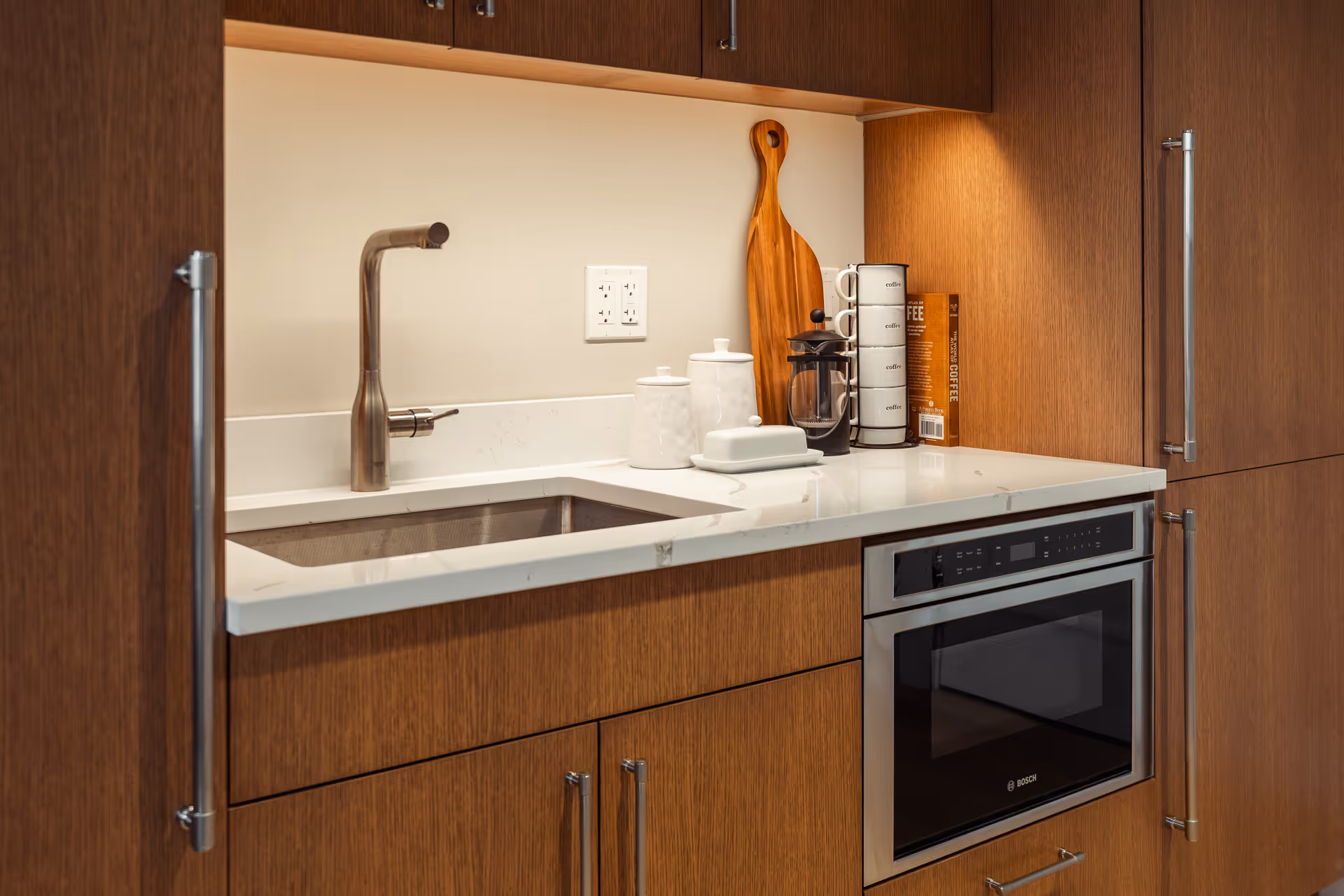 A modern kitchen area with wooden cabinets, a white countertop, a built-in stainless steel Bosch microwave oven, a stainless steel faucet over a sink, and kitchen accessories including a wooden cutting board, a French press, stacked coffee mugs, and canisters.