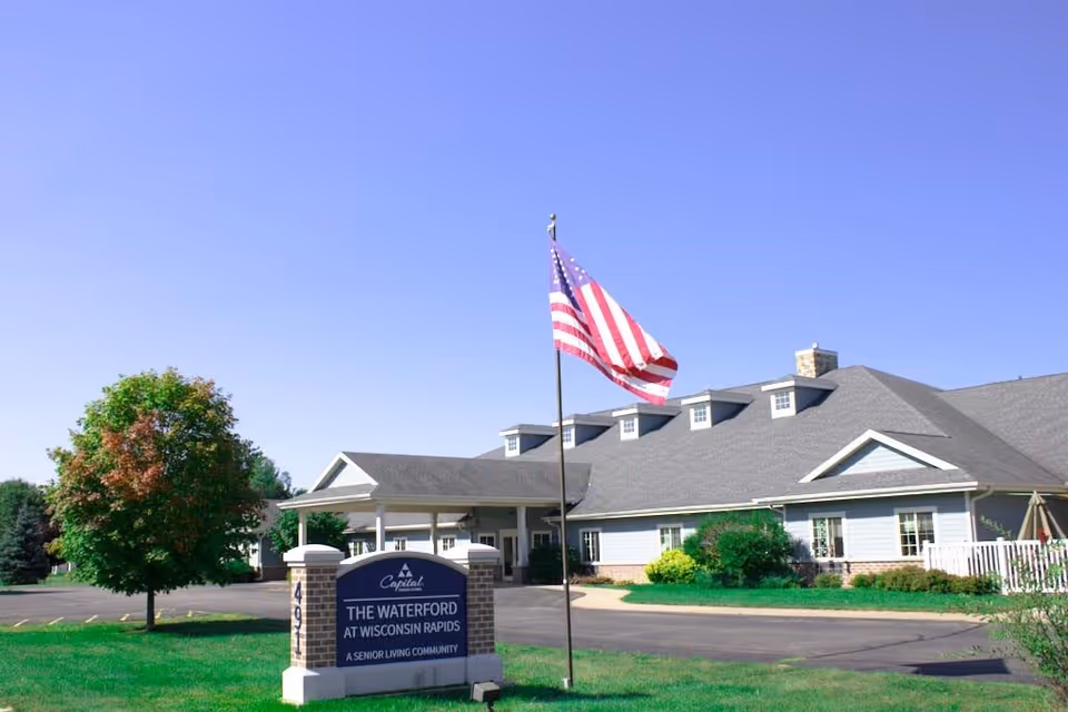 Exterior view of The Waterford at Wisconsin Rapids senior living community building with a large American flag on a flagpole in front and a sign displaying the facility name. The building has a gray roof, light-colored siding, and is surrounded by green grass and trees under a clear blue sky.