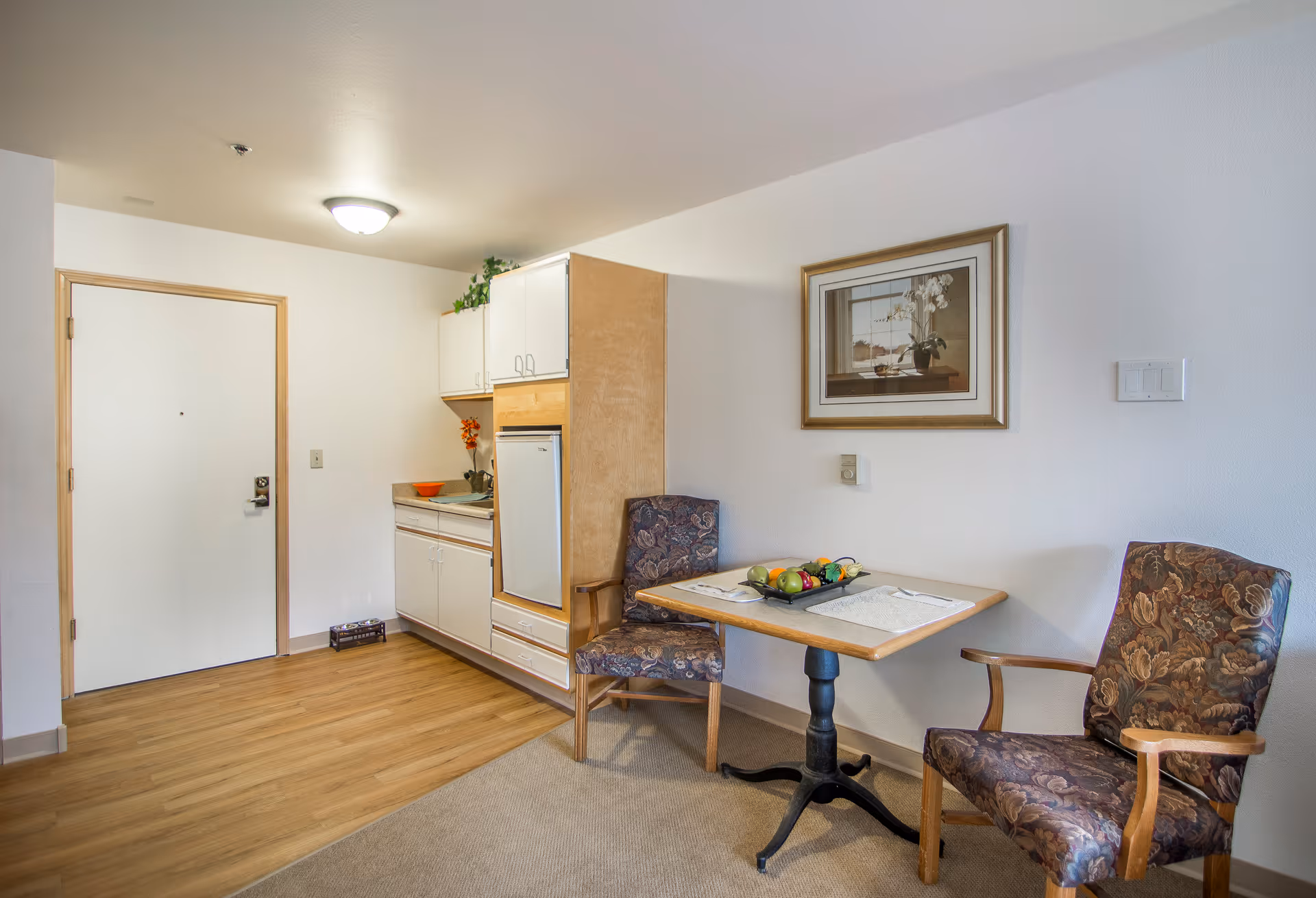 A small dining area in a senior living facility with a wooden table set for two, featuring a decorative fruit centerpiece. Two upholstered chairs with floral patterns are placed around the table. To the left, there is a kitchenette with white cabinets, a small refrigerator, and a countertop with a sink. The room has light-colored walls, a wooden floor near the kitchenette, and carpet near the dining area. A framed picture hangs on the wall above the table.