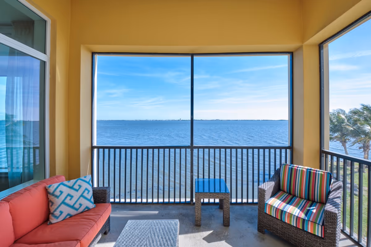 A screened balcony with a view of a large body of water and a clear blue sky. The balcony has a red cushioned sofa with a patterned pillow, a wicker armchair with a colorful striped cushion, and a small wicker table. Palm trees are visible to the right outside the balcony.