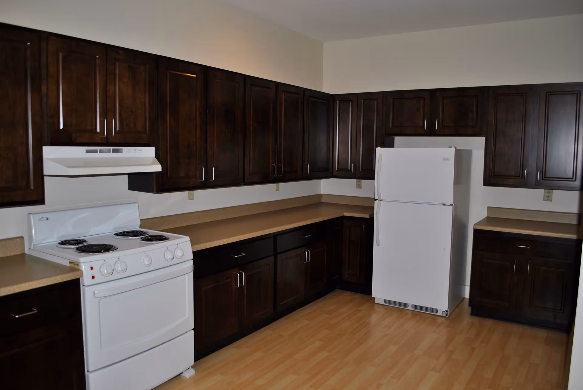 A kitchen with dark wooden cabinets, a white electric stove with four coil burners and an oven, a white refrigerator, beige countertops, and light wood flooring.