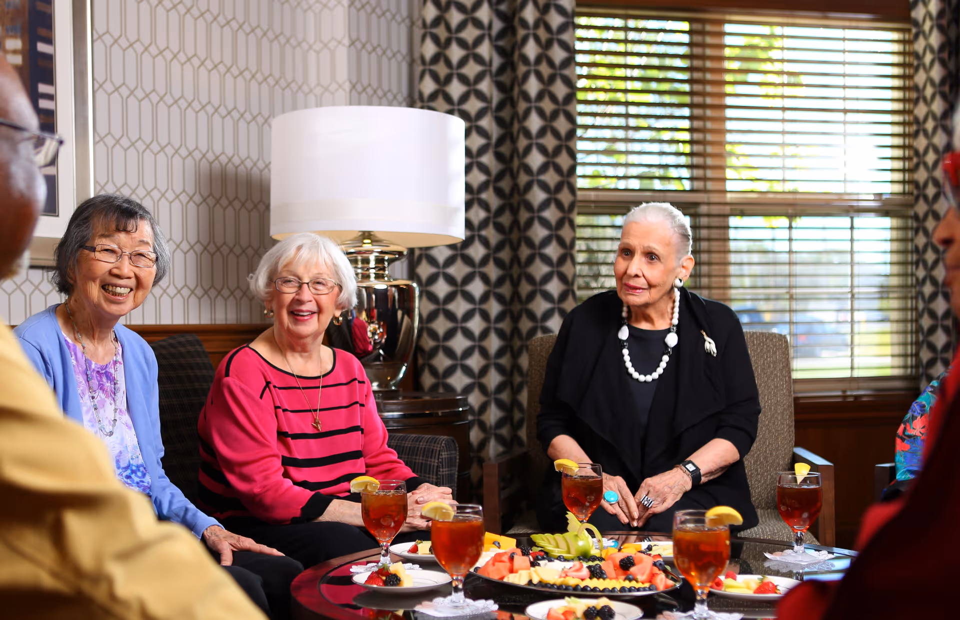 Three elderly women seated in a communal living room smiling around a table with iced tea and a fruit platter.