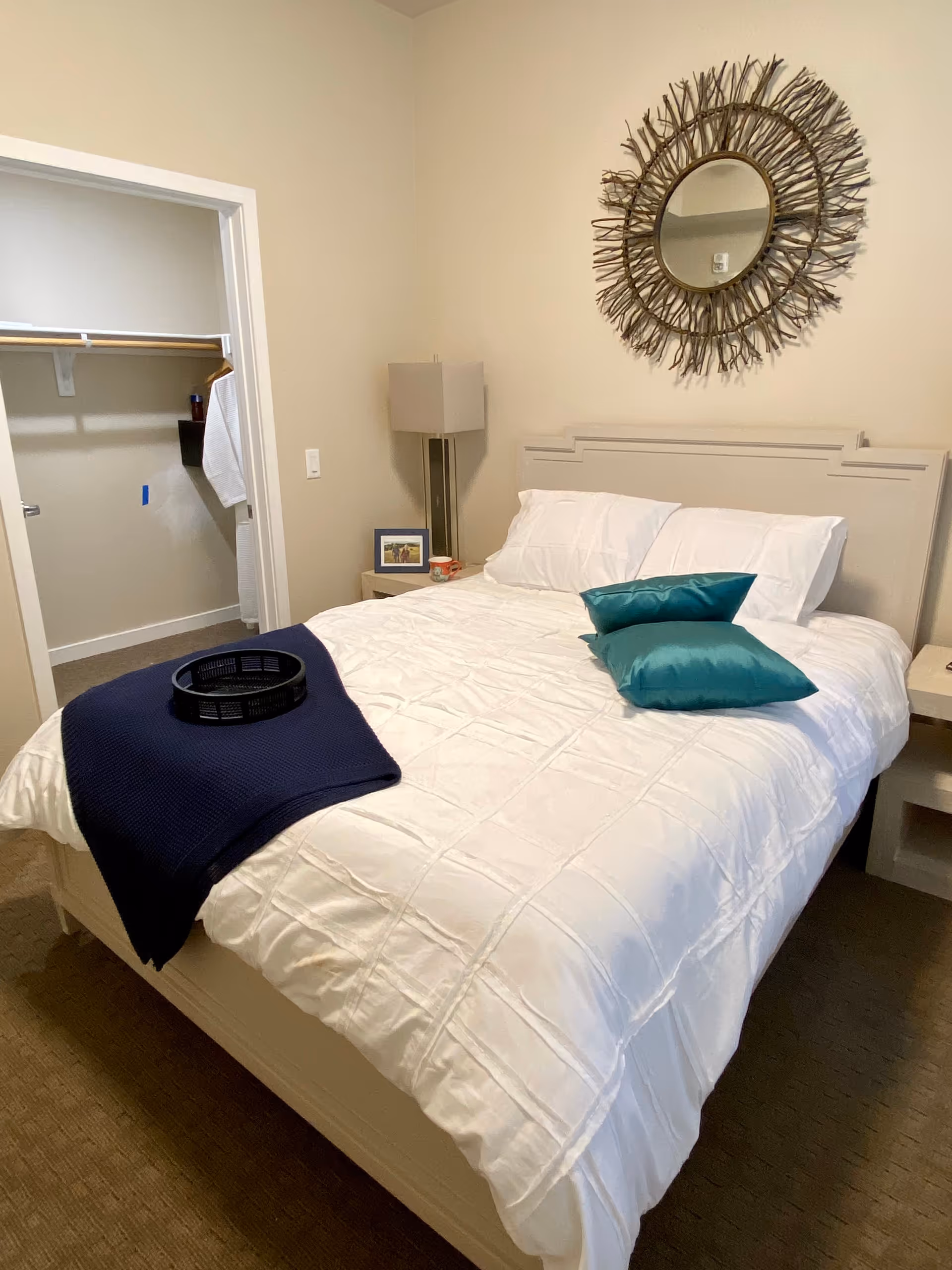 A bedroom with a neatly made bed featuring white bedding and two teal pillows. A dark blue throw blanket and a black tray are placed on the bed. There is a nightstand with a lamp, a framed photo, and a mug. A decorative round mirror with a twig-like frame hangs on the beige wall above the bed. An open walk-in closet is visible to the left.