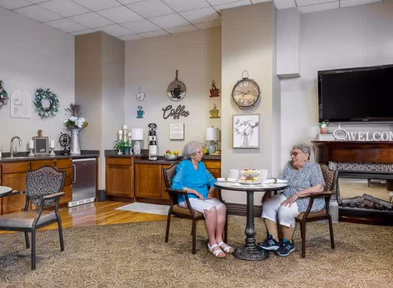 Two elderly women sitting at a round table in a cozy common area with a coffee station, decorative wall art, and a fireplace with a TV above it. The room has wooden cabinets, a patterned carpet, and comfortable chairs.