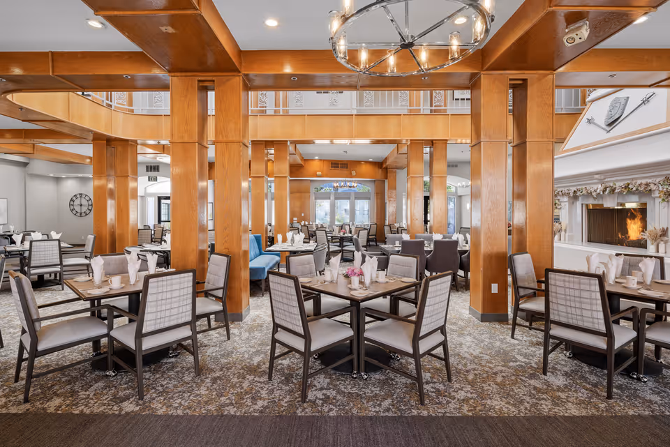 A spacious dining room in a senior living community featuring multiple tables set with white napkins, cups, and plates. The room has wooden pillars and beams, a patterned carpet, and a cozy fireplace decorated with garlands. There are cushioned chairs around the tables and a chandelier hanging from the ceiling.