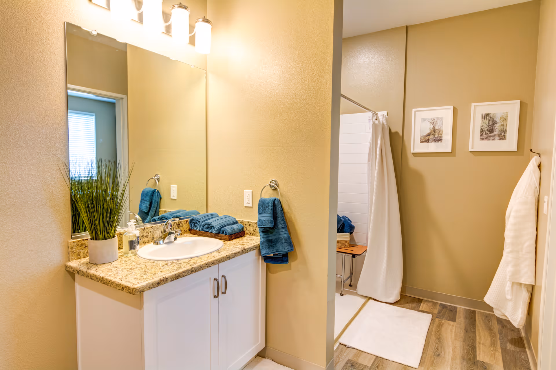 A clean and well-lit bathroom with a granite countertop sink, a large mirror, and three light fixtures above. Blue towels are neatly folded on the counter and hanging on a towel ring. There is a potted plant and a soap dispenser on the counter. In the background, a shower area with a white curtain and a small wooden bench is visible. Two framed pictures hang on the wall, and a white bathrobe is hanging on a hook on the right wall. The floor has wood-like tiles and a white bath mat.