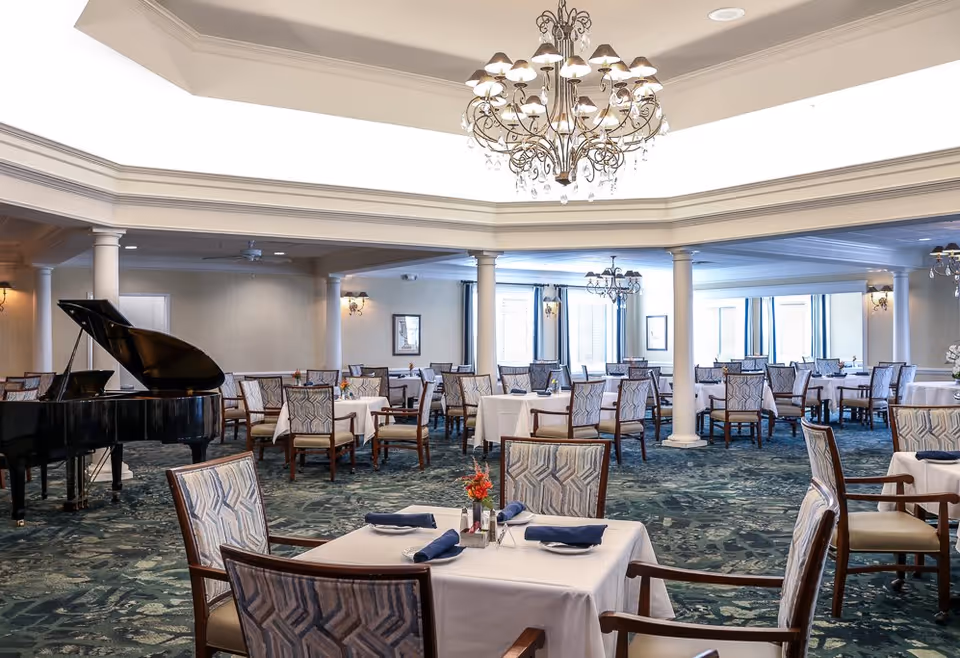 A spacious dining room with multiple tables covered in white tablecloths, each set with blue napkins and small floral centerpieces. The room features patterned chairs, a grand piano on the left side, elegant chandeliers hanging from the ceiling, and large windows allowing natural light to fill the space.