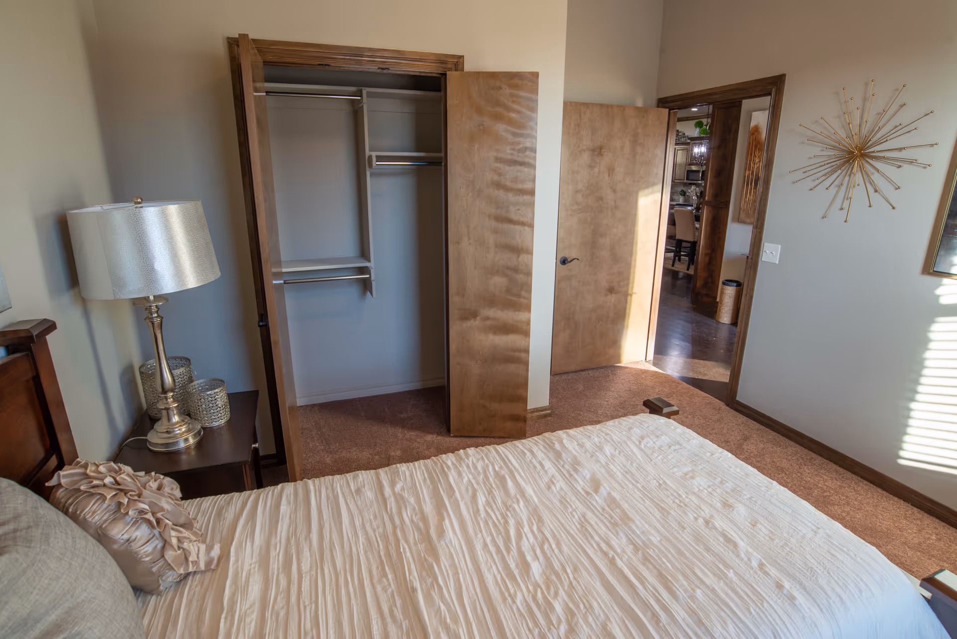A bedroom with a bed covered in a beige textured bedspread and a decorative pillow. To the left of the bed is a wooden nightstand with a silver lamp and decorative items. The closet door is open, showing empty shelves and hanging rods. The bedroom door is also open, revealing a view into another room with wooden flooring and dining chairs. The walls are painted light beige, and there is a sunburst wall decoration on the right wall.