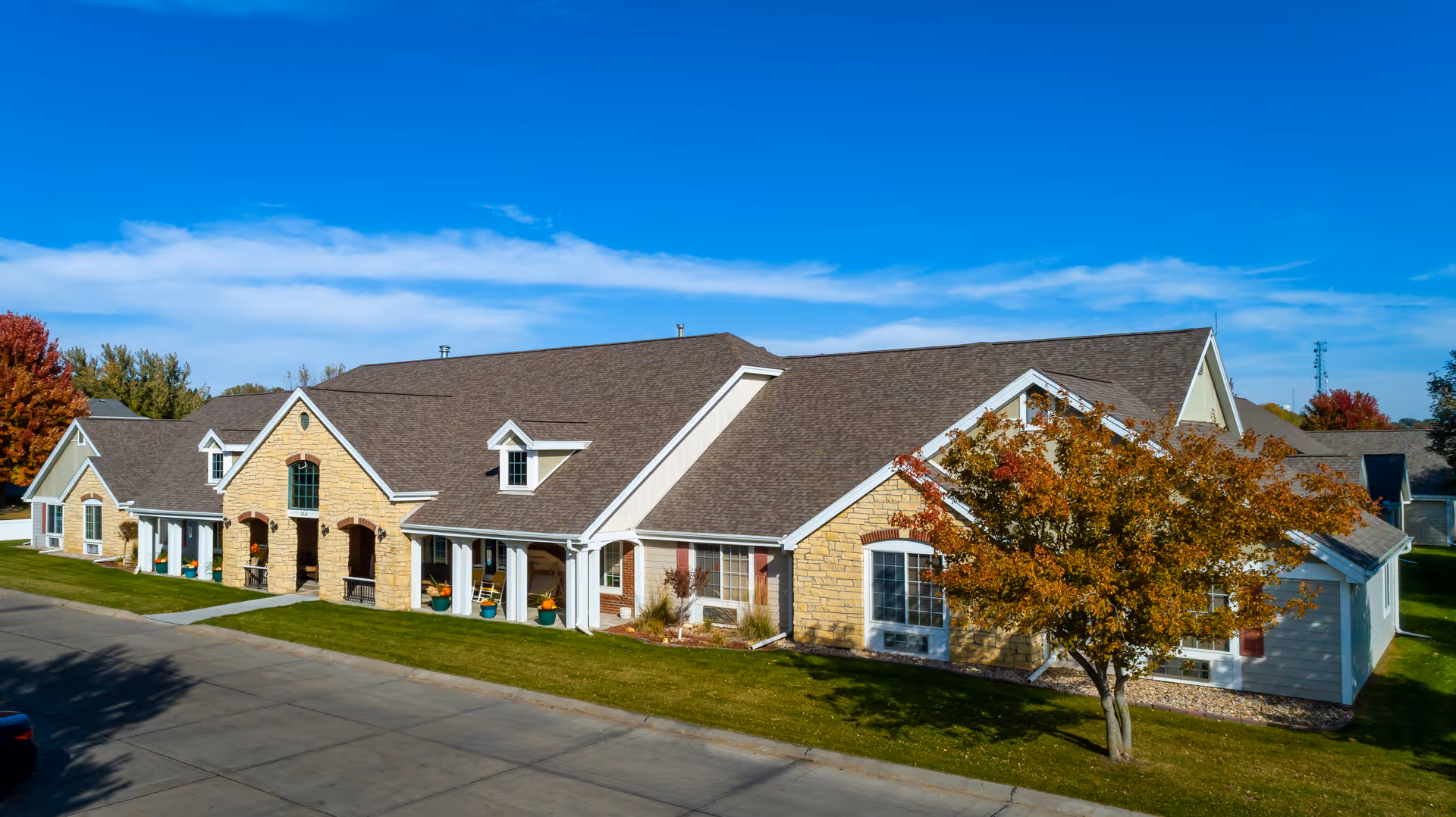 Exterior view of a single-story assisted living facility building with a stone and siding facade, multiple windows, and a sloped roof. There is a tree with autumn-colored leaves in front of the building, a well-maintained lawn, and a clear blue sky above.