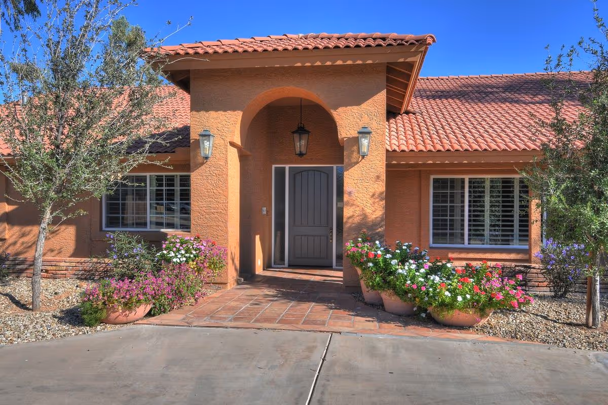 Front entrance of a single-story Mediterranean-style building with a tiled roof, arched doorway, and potted flowering plants.