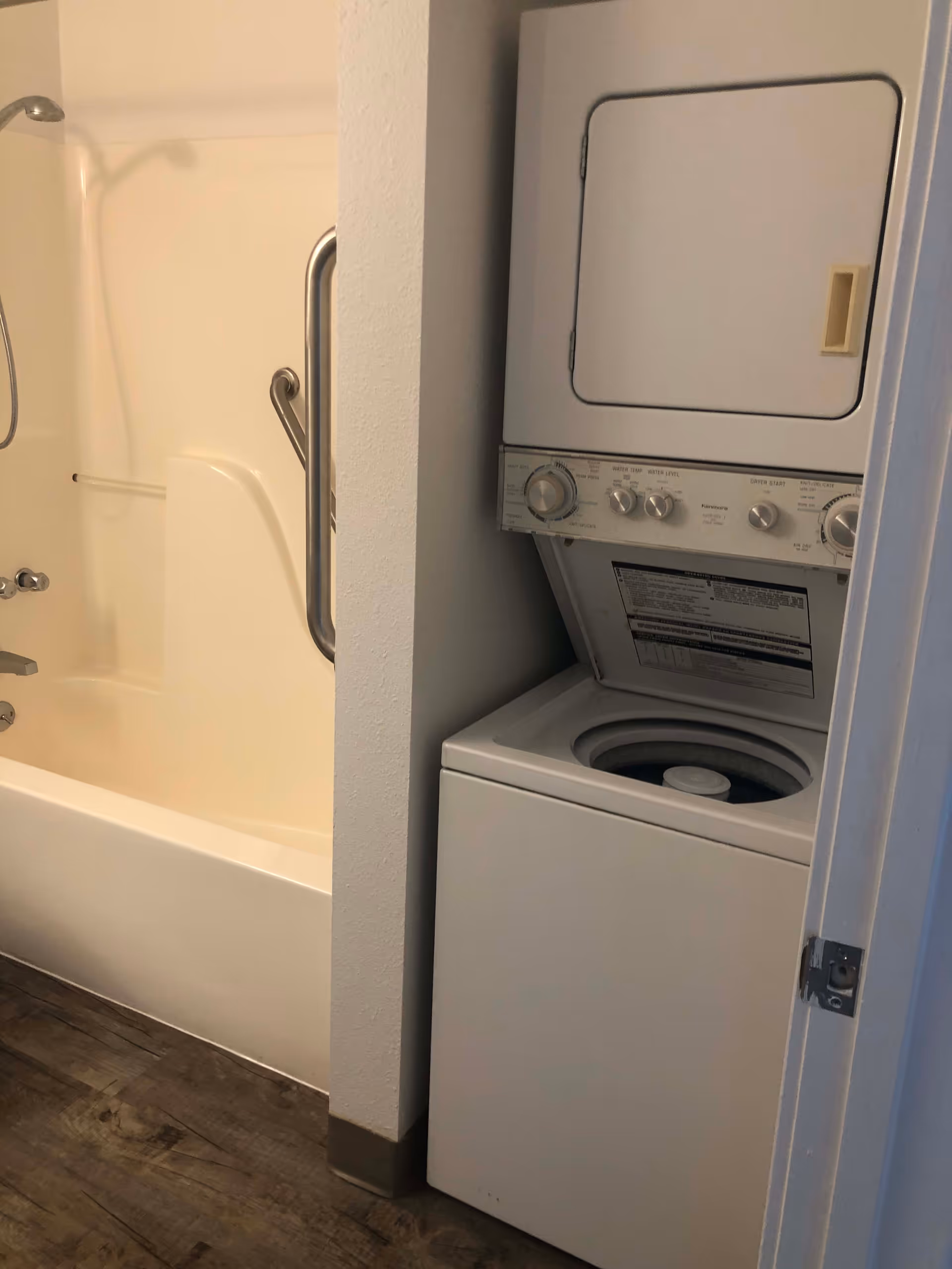 A small laundry area with a stacked washer and dryer unit next to a bathtub with a grab bar, inside a senior living facility.