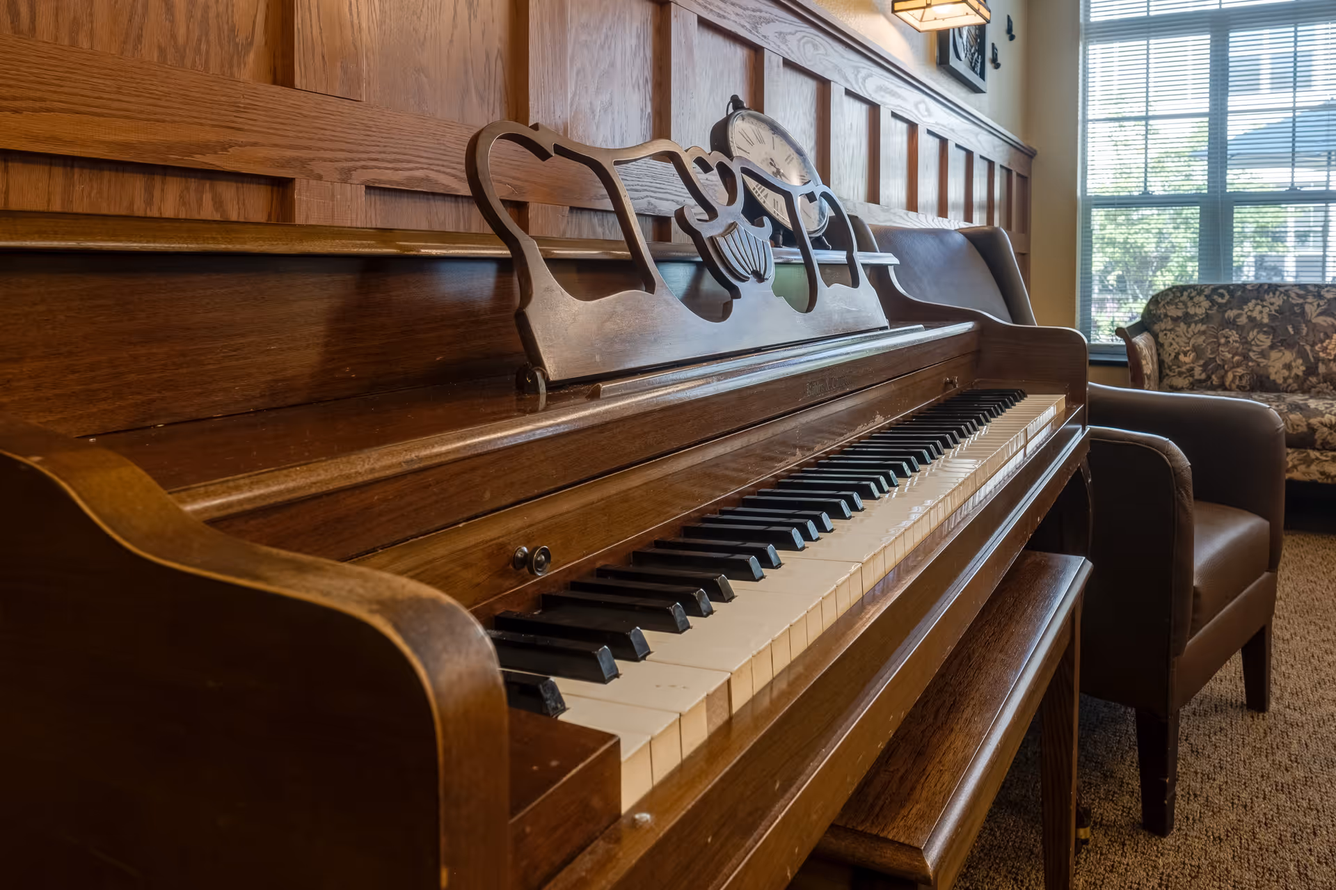 Close-up view of a wooden upright piano with a decorative music stand in a room featuring wood paneling on the wall, a brown armchair, a floral-patterned sofa, and a window with blinds letting in natural light.