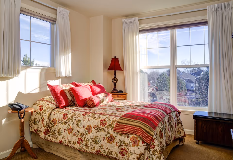 Sunlit bedroom with a floral bedspread, red pillows, a bedside lamp, and large windows overlooking a neighborhood.