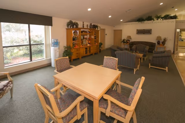 A spacious common area in a senior living facility featuring a square wooden table with four cushioned chairs in the foreground. In the background, there are several upholstered armchairs and sofas arranged around small tables. A wooden cabinet with decorative items and plants is against the wall, and a large window lets in natural light. The room has carpeted floors and neutral-colored walls with ceiling lights.
