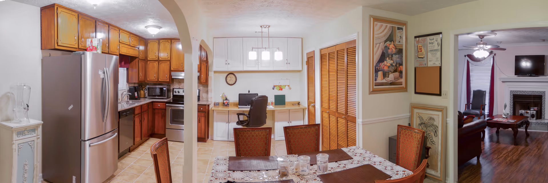 Interior view of a residential home showing a kitchen with wooden cabinets, stainless steel refrigerator, stove, and microwave on the left. In the center is a dining area with a table covered by a white and floral tablecloth and four chairs. Behind the dining area is a small office nook with a desk, chair, computer, and white cabinets above. On the right side is a living room with a fireplace, TV mounted above it, a ceiling fan, and seating furniture. The floors are tiled in the kitchen and dining area, and wood in the living room.