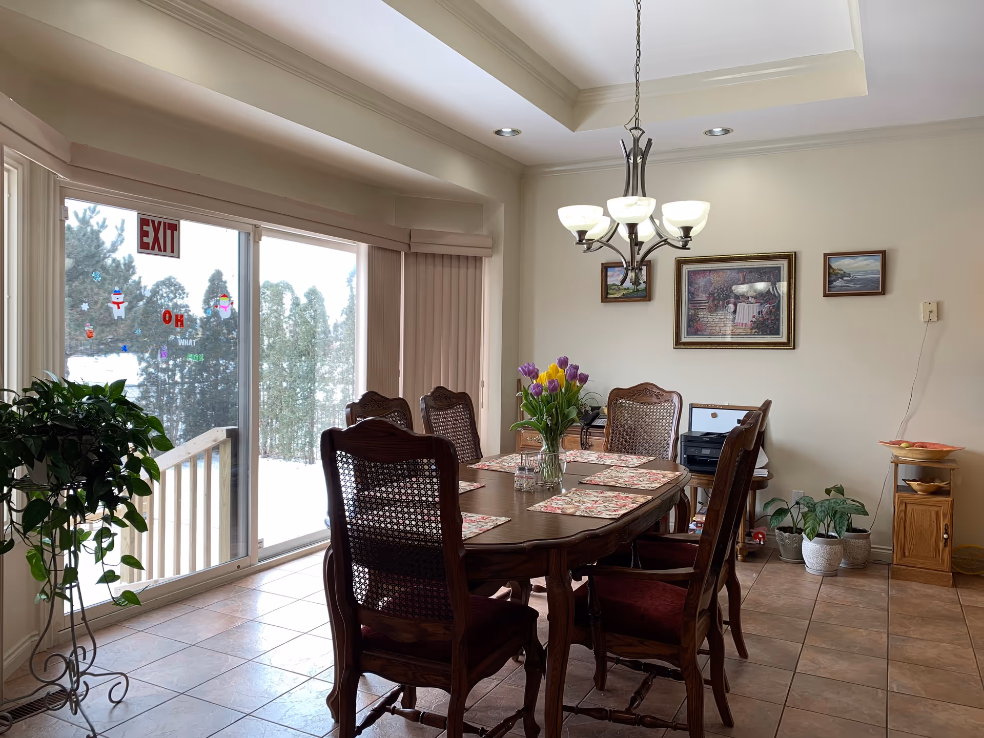 A dining room with a wooden table and six chairs. The table has floral placemats and a vase with colorful tulips. There are three framed pictures on the wall, a chandelier hanging from the ceiling, and several potted plants around the room. Large sliding glass doors with an exit sign lead outside, showing snow and trees.