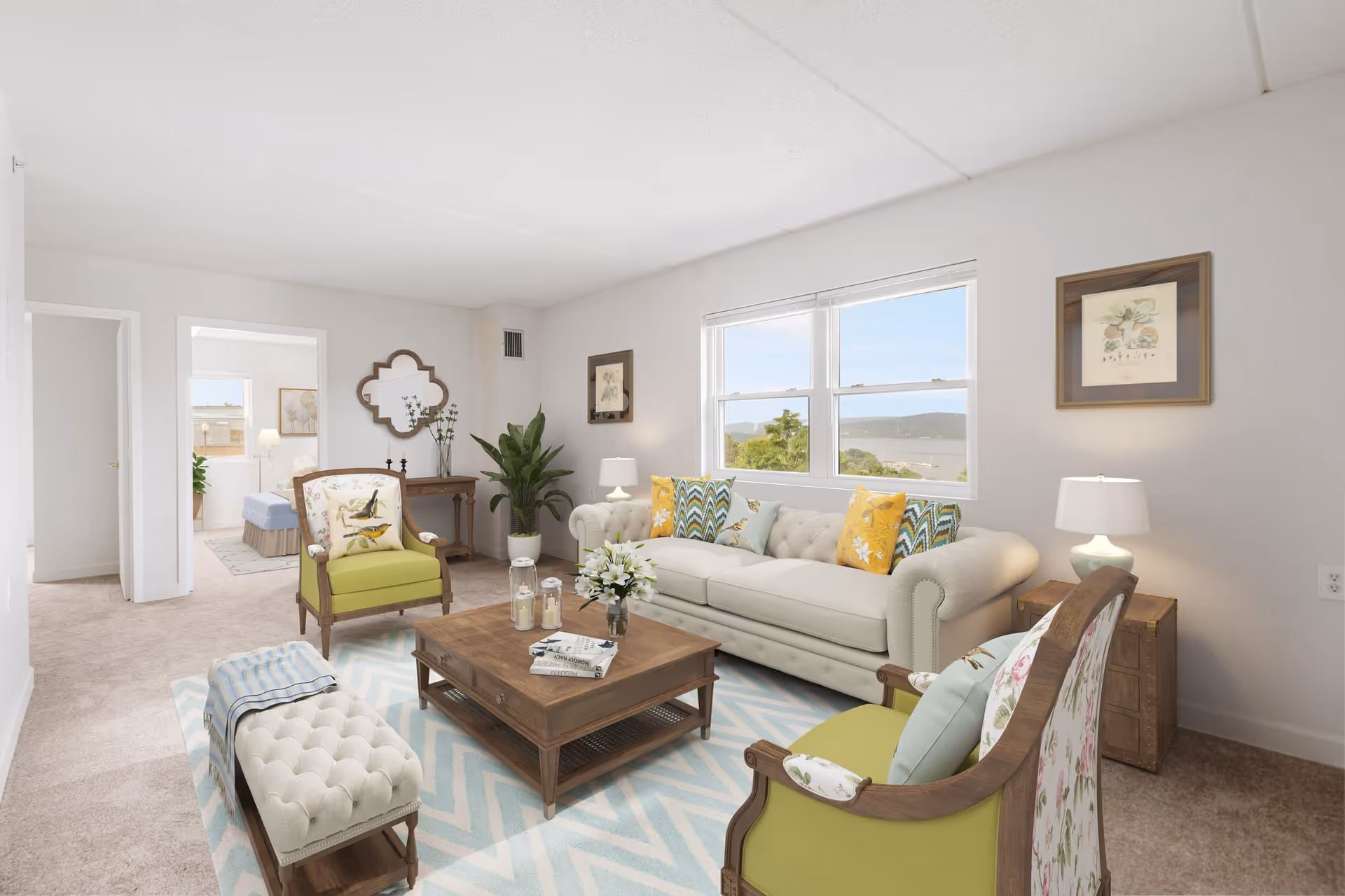 A bright and cozy living room in a senior living community featuring a beige tufted sofa with colorful patterned pillows, two green armchairs with floral cushions, a wooden coffee table with books and candles, a cushioned bench with a blue striped throw, a potted plant, framed artwork on the walls, and a large window showing a scenic view of trees and hills outside.