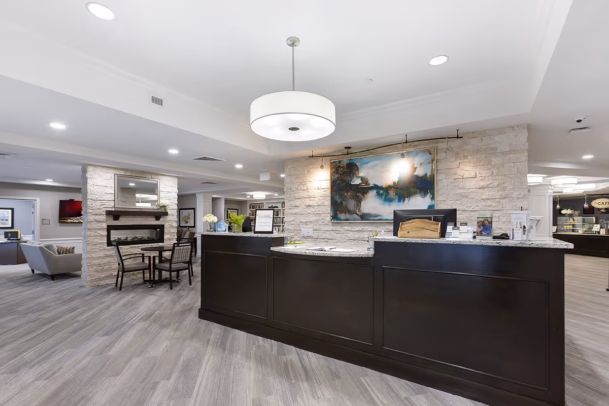 Reception area of StoryPoint Medina featuring a dark wood front desk with a granite countertop, a large modern pendant light hanging from the ceiling, a stone accent wall with a framed abstract painting, and seating areas with tables and chairs. The space is well-lit with recessed lighting and has a cozy fireplace in the background.