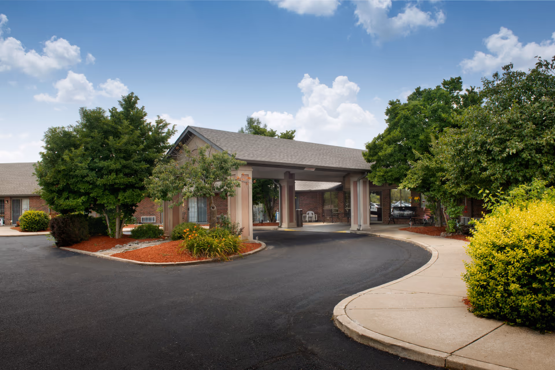 Entrance driveway of a senior living facility with a covered drop-off area, surrounded by landscaped greenery and trees under a partly cloudy sky.