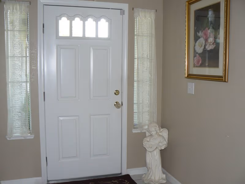 Interior view of an entryway featuring a white front door with decorative glass panels at the top, flanked by two narrow windows with sheer curtains. To the right of the door, there is a small statue of an angel holding a musical instrument and a framed floral painting on the beige wall.