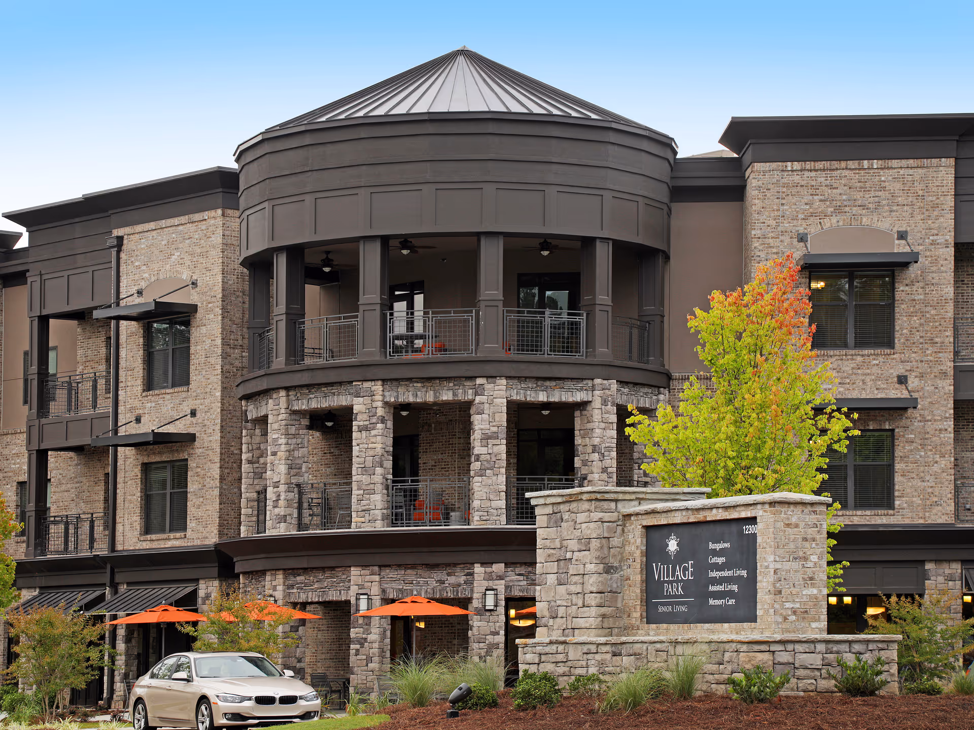 Exterior view of Village Park Alpharetta senior living facility showing a multi-story building with stone and brick facade, balconies, and a prominent round turret-like structure. There are orange umbrellas on the patio area and a stone sign in front displaying the facility name and services offered.
