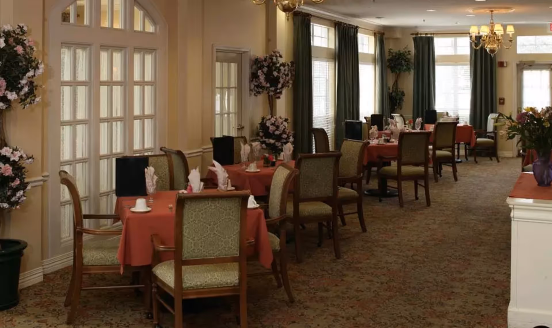 A dining room with several tables covered in red tablecloths, each set with folded napkins, cups, and small flower arrangements. The room has large windows with green curtains, floral decorations, and a chandelier hanging from the ceiling. The carpet has a patterned design, and the walls are painted a light beige color.