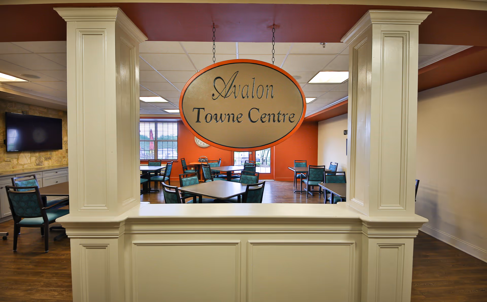 Interior view of a common area named Avalon Towne Centre with several tables and chairs arranged for seating. The room has a warm color scheme with orange and beige walls, wood flooring, and a large window letting in natural light. A flat-screen TV is mounted on a stone accent wall to the left.