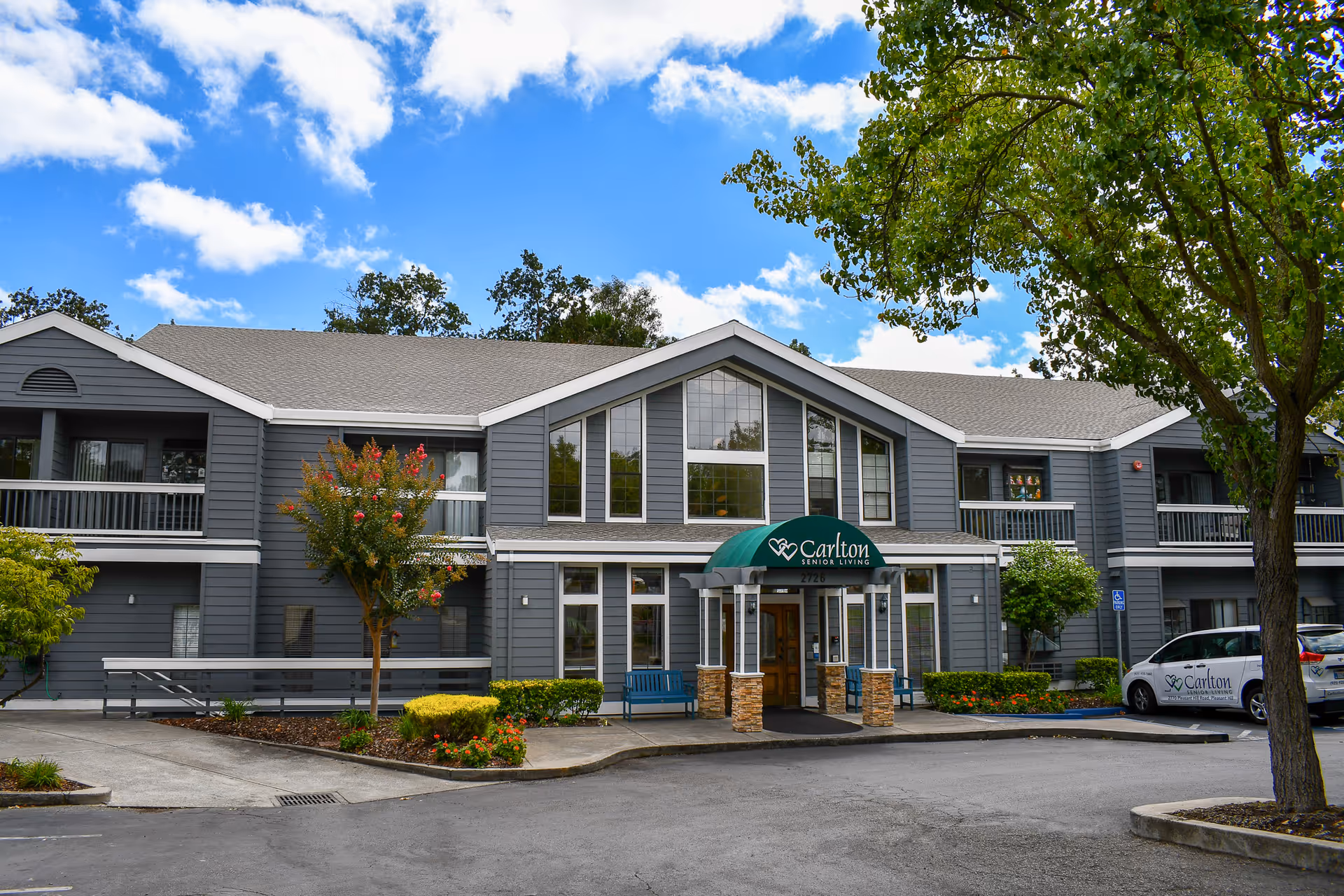 A gray two-story senior living facility with a green awning entrance and landscaped surroundings.