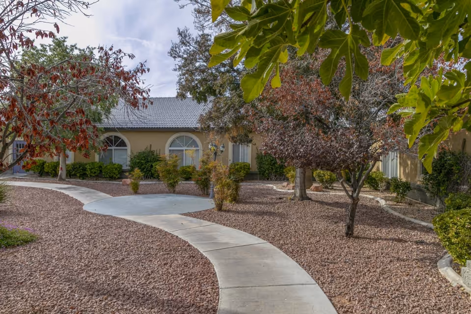 Curved concrete pathway leading through a landscaped garden area with trees and shrubs in front of a single-story building with arched windows and a tiled roof under a partly cloudy sky.