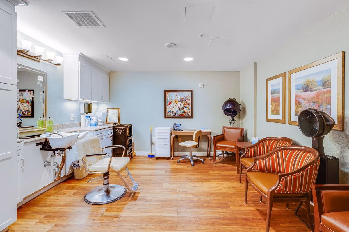Interior view of a salon area in a senior living facility with a hair washing station, salon chair, wooden floor, and several seating chairs. The walls are decorated with framed floral artwork, and there is a desk with a chair and hair drying equipment in the corner.